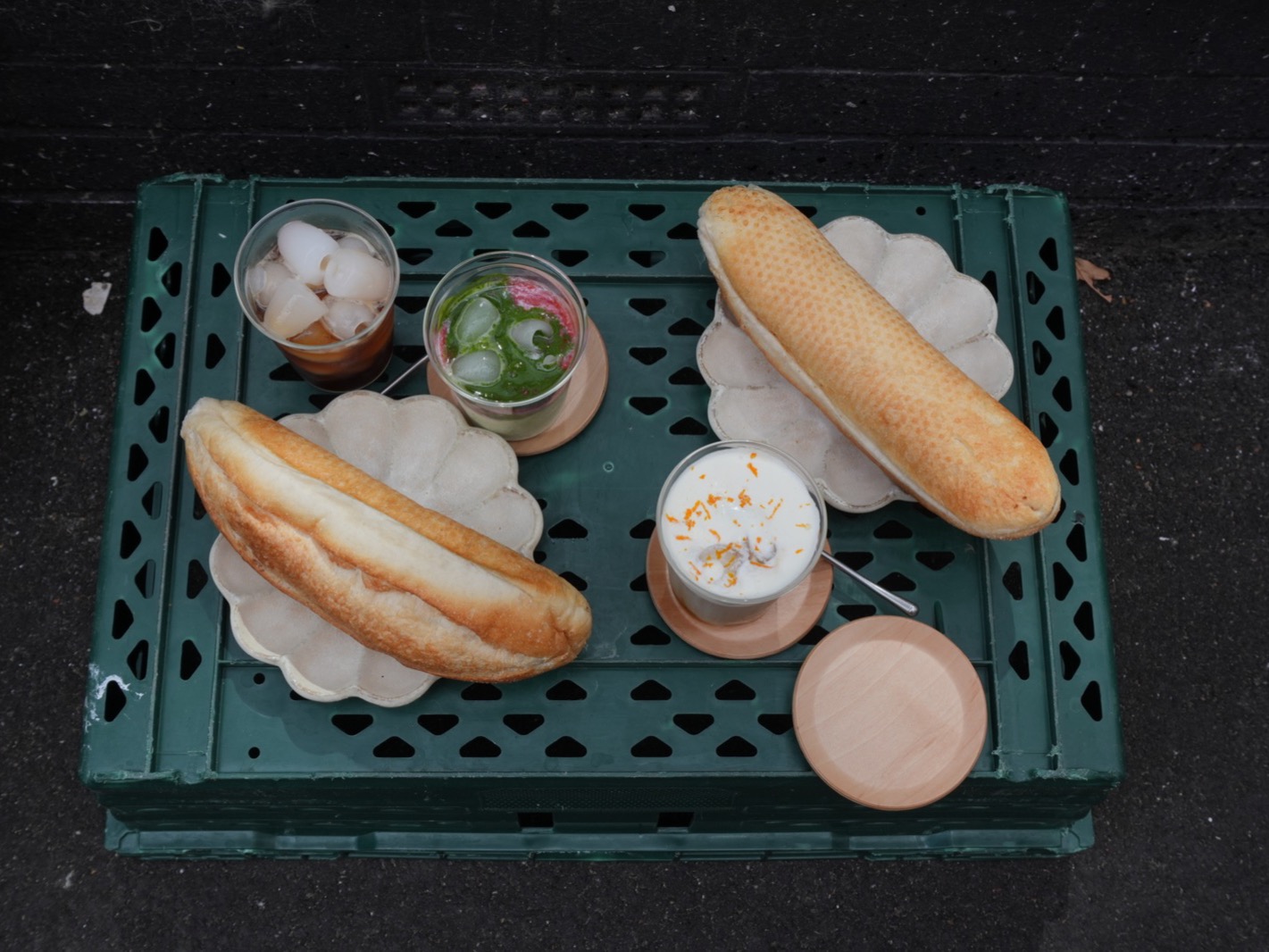 Overhead view of a green plastic crate used as a table: two bánh mì baguettes on scalloped plates, a glass of ice, a matcha drink and a coffee topped with orange zest.