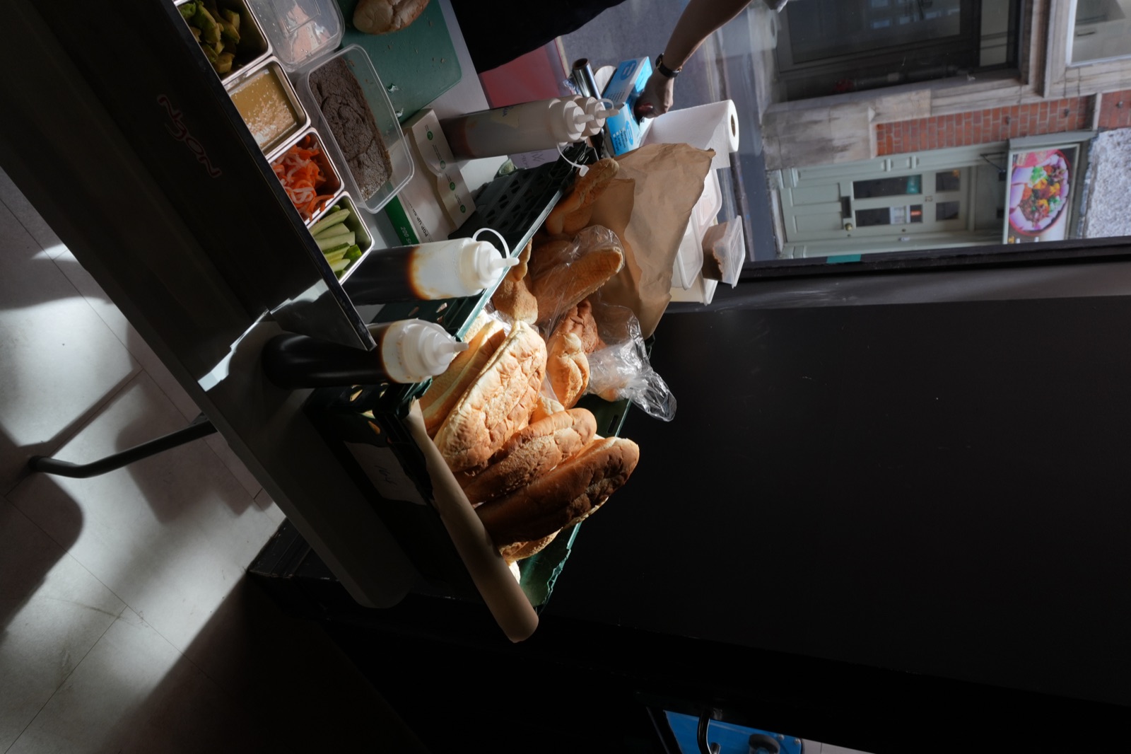 The NgON prep station set up beside a shop window: a green crate of baguettes, squeeze bottles of sauce and trays of pâté, pickled carrot and cucumber, with the street visible outside.